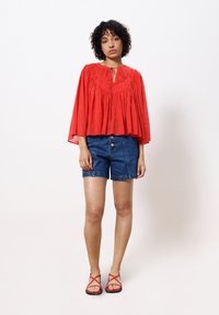 Rote, lässig sitzende Bluse mit Raffung und Bindeband, kombiniert mit blauen Jeansshorts und roten Riemchensandalen.