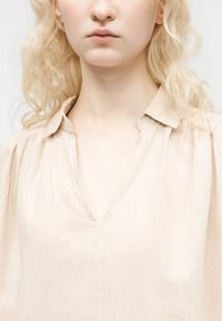 Ξανθός με χοντρά, μαλακά σγουρά μαλλιά που φοράει μπεζ μπλούζα με V λαιμόκοψη και γιακά, εμφανιζόμενος από τους ώμους μέχρι το μέσο του στήθους.