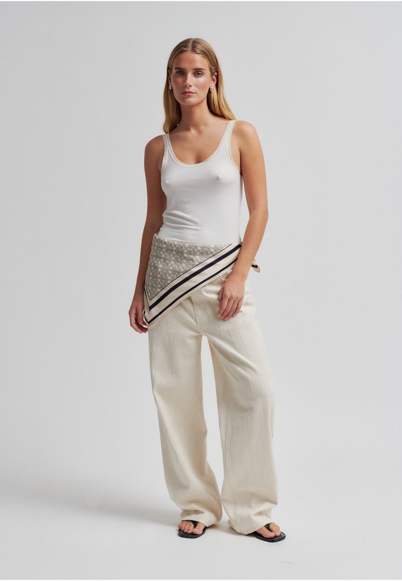 Femme portant un haut blanc sans manches, un pantalon beige à jambes larges, des sandales noires et une écharpe à motifs attachée autour de la taille, debout devant un fond uni.