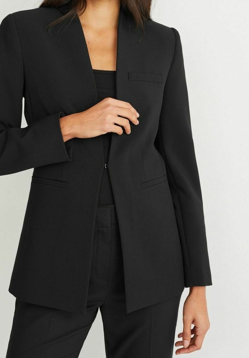 Femme portant un blazer noir sur mesure par-dessus un top noir, avec un pantalon noir assorti, ajustant le devant du blazer.