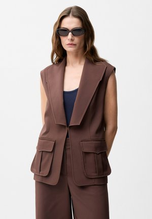 Vrouw draagt een mouwloos bruin op maat gemaakt vest met grote revers, bijpassende bruine broek, donkerblauwe top en zwarte zonnebril.