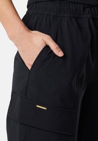 Musta cargo-housut, joissa on joustava vyötärö, sivutasku ja kultainen brändilogo-yksityiskohta. Valmistettu sileästä kankaasta, rento istuvuus.