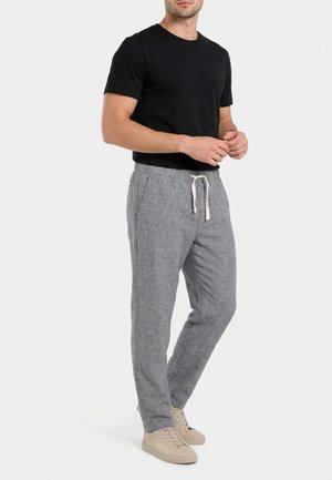 Homme portant un t-shirt noir à manches courtes, un pantalon gris à cordon et des baskets beiges, se tenant devant un fond blanc uni.