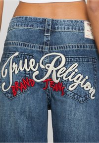 Džínsy s tmavomodrým prácovaním, s vyšívaným nápisom "True Religion" v bielej a červenej farbe na zadných vreckách, so štandardným dizajnom vreciek.