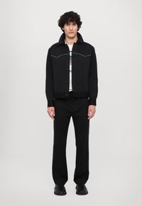 Černá zkrácená bunda s zeleným prošíváním, zapínání na knoflíky a vysokým límcem, v kombinaci s černými kalhotami se bočními pruhy a teniskami.