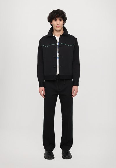 Black cropped jacket with green stitching, button closure, and high collar, paired with black pants featuring side stripes and sneakers.
