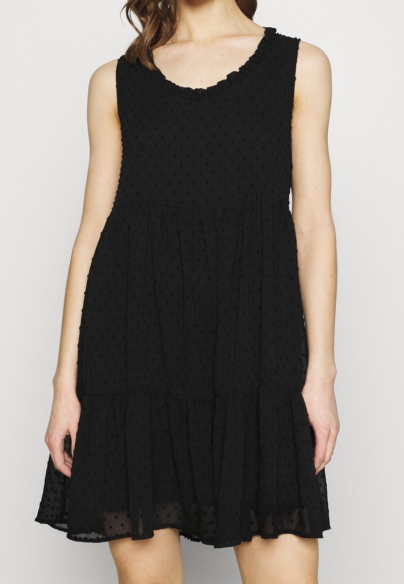 Robe noire texturée sans manches avec encolure américaine à volants et jupe à étages, portée par une femme debout sur un fond neutre.