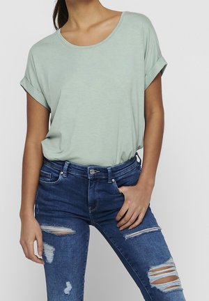 Mujer con camiseta de manga corta verde claro metida en vaqueros ajustados azules rotos, con una mano en el bolsillo, frente a un fondo liso.
