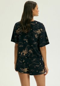 Kobieta w czarnym piżamowym zestawie z beżowymi wzorami przedstawiającymi Pegaza, półksiężyc, gwiazdy i chmurki, widoczna z tyłu na jednolitym tle.