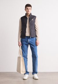 Gilet nero imbottito con trapuntatura verticale, maglione beige chiaro, jeans blu e sneakers bianche. Tiene una borsa a tracolla beige.