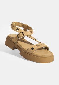Beige Plateausandalen mit einem breiten Riemen, der runde metallische Akzente aufweist, und verstellbaren Knöchelriemen. Strukturiertes Sohlenmaterial mit Zickzackmuster.