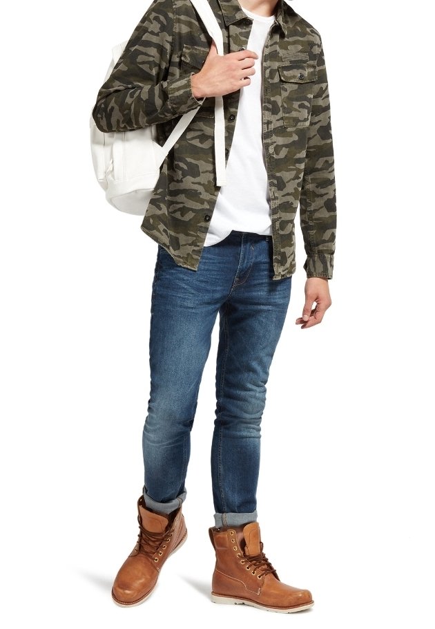 Camicia a bottoni camouflage, t-shirt bianca, jeans skinny blu, stivali marroni con lacci. Gli accessori includono uno zaino bianco.