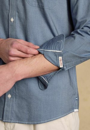 Mann krempelt den Ärmel eines hellblauen Hemdes mit Knöpfen hoch, zeigt die weiße Innenseite der Manschette und ein kleines Stoffetikett.