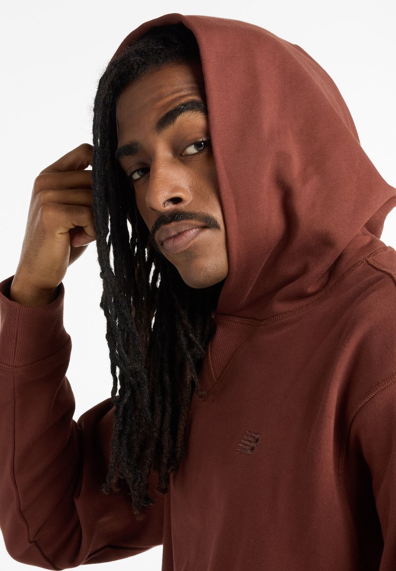 Brown hoodie with a large hood, made of soft fabric. Features ribbed cuffs and a subtle logo on the chest. Model shows a side profile.