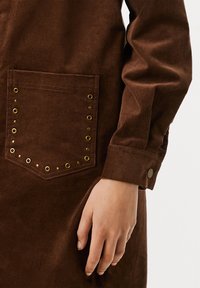 Burgundijas korduroja krekls ar zeltainiem piespraužamiem kabatām un pogādām. Gluda struktūra ar taisnu griezumu.