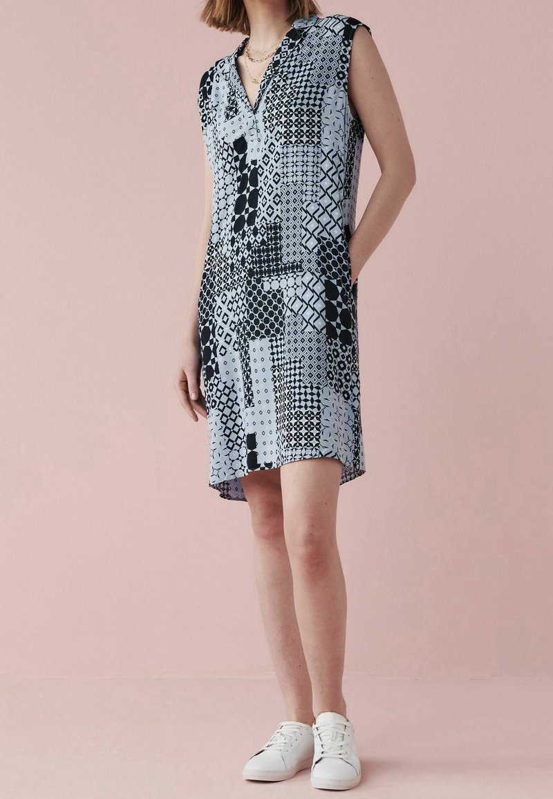 Femme portant une robe sans manches mi-longue avec des motifs géométriques noirs et blancs et des baskets blanches, debout devant un fond rose.