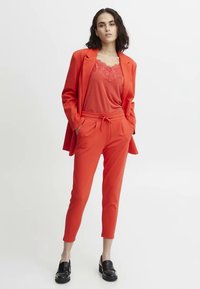 Fato vermelho com um casaco estruturado e calças justas, combinado com uma blusa sem mangas com detalhes em renda e sapatos loafers pretos.