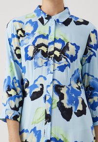 Květinová košile na knoflíky z lehké látky. Má velké květinové vzory v modré, černé a zelené barvě, s světle modrým pozadím a límcem.
