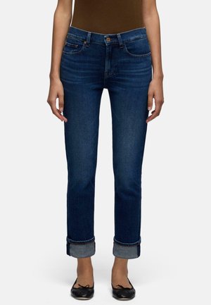 Frau trägt dunkelblaue umgekrempelte Jeans und schwarze Ballerinas, gezeigt von der Taille bis zu den Füßen vor einfachem Hintergrund.