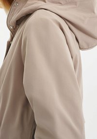 Světlý béžový kabát s kapucí a hladkou matnou texturou látky, s kovovými patentními knoflíky a zipem podél přední části.