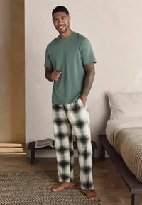 T-shirt en coton vert et pantalon de pyjama à carreaux crème avec des accents noirs. Coupe décontractée. Debout sur un tapis tissé, souriant à la caméra.