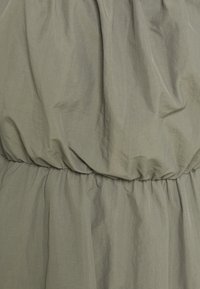 Robe vert olive avec taille froncé et tissu fluide. La texture est lisse et légèrement froissée, présentant des panneaux élastiques à la taille.
