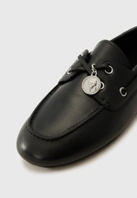 Schwarzer Lederschuh mit runder Zehenform, ausgestattet mit silbernen Ösen, einem geschnürten Design und einem runden Logo-Anhänger, der an den Schnürsenkeln befestigt ist.