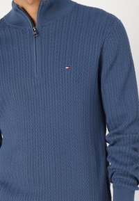 Sininen neulepaita, jossa on teksturoitu timanttikuvio, neljännesvetoketjukaulus ja pieni brodeerattu logo punaisena, valkoisena ja sinisenä rinnassa.