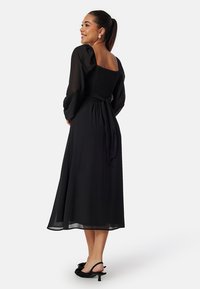 Abito maxi nero con maniche a sbuffo e corpetto arricciato. Tessuto morbido, cintura in vita e gonna fluida. Scarpe nere aperte davanti.