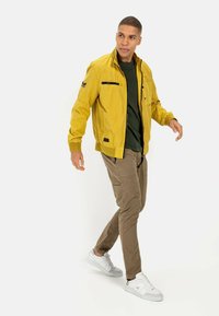 Gelbe zip-up Jacke mit mehreren schwarzen Akzenten und Taschen, getragen über einem dunkelgrünen Shirt, kombiniert mit beigen Cargo-Hosen und weißen Sneakern.