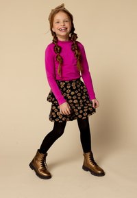 Meisje met gevlochten haar en een lichtbruine haarband draagt een fuchsia lange mouwen top, een zwarte bloemenrok, zwarte panty’s en metallic bronzen laarzen, en glimlacht.