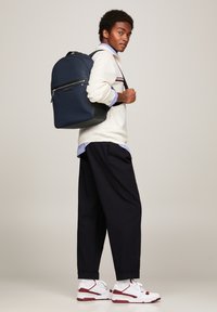 Zaino navy dal design elegante, dotato di una tasca frontale con zip e forma strutturata, abbinato a un maglione crema e pantaloni scuri.