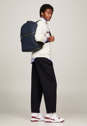 Zaino navy dal design elegante, dotato di una tasca frontale con zip e forma strutturata, abbinato a un maglione crema e pantaloni scuri.