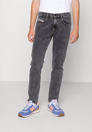 Personne portant un jean noir délavé et des baskets bleues New Balance, debout devant un fond gris clair uni.