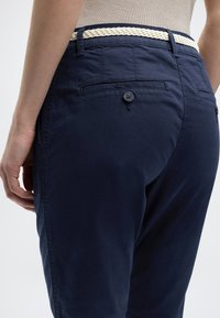 Άτομο που φοράει μπλε σκούρο παντελόνι με τσέπη στο πίσω μέρος κλειστή με κουμπί και λευκή πλεκτή ζώνη, στέκεται με το ένα χέρι χαλαρό στο πλάι.