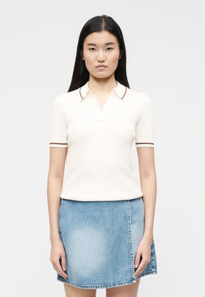 Nuori nainen, jolla on pitkät tummat hiukset, valkoinen lyhythihainen poolopaita, jossa ruskea reunus, ja sininen farkkuhame, seisoo yksiväristä taustaa vasten.