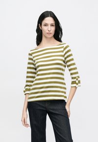 Svītrains garroku tops olīvu zaļā un baltā krāsā ar plašu kakla izgriezumu; aprīkots ar sarullētiem piedurknēm un pogu akcentiem. Ikdienišķa piegriezuma.