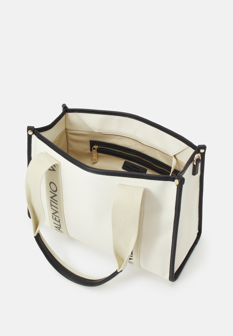 Bolso de lona en color crema con ribetes negros, que cuenta con dos asas, un bolsillo con cremallera en el interior y una etiqueta de marca visible en el lateral.