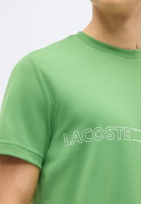 Κοντινό πλάνο ενός ατόμου που φοράει πράσινο κοντομάνικο πουκάμισο με λευκό περίγραμμα κειμένου "LACOSTE" στο στήθος.