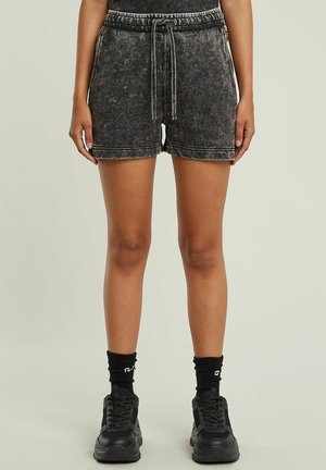 Personne debout portant un short noir délavé à l'acide avec cordon, des chaussettes noires aux chevilles et des baskets noires épaisses sur un fond uni.