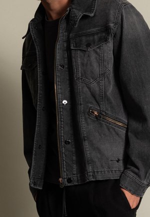 Hombre con chaqueta de denim gris oscuro, con bolsillos con cremallera y botones, camiseta negra debajo y manos en los bolsillos de los pantalones negros.