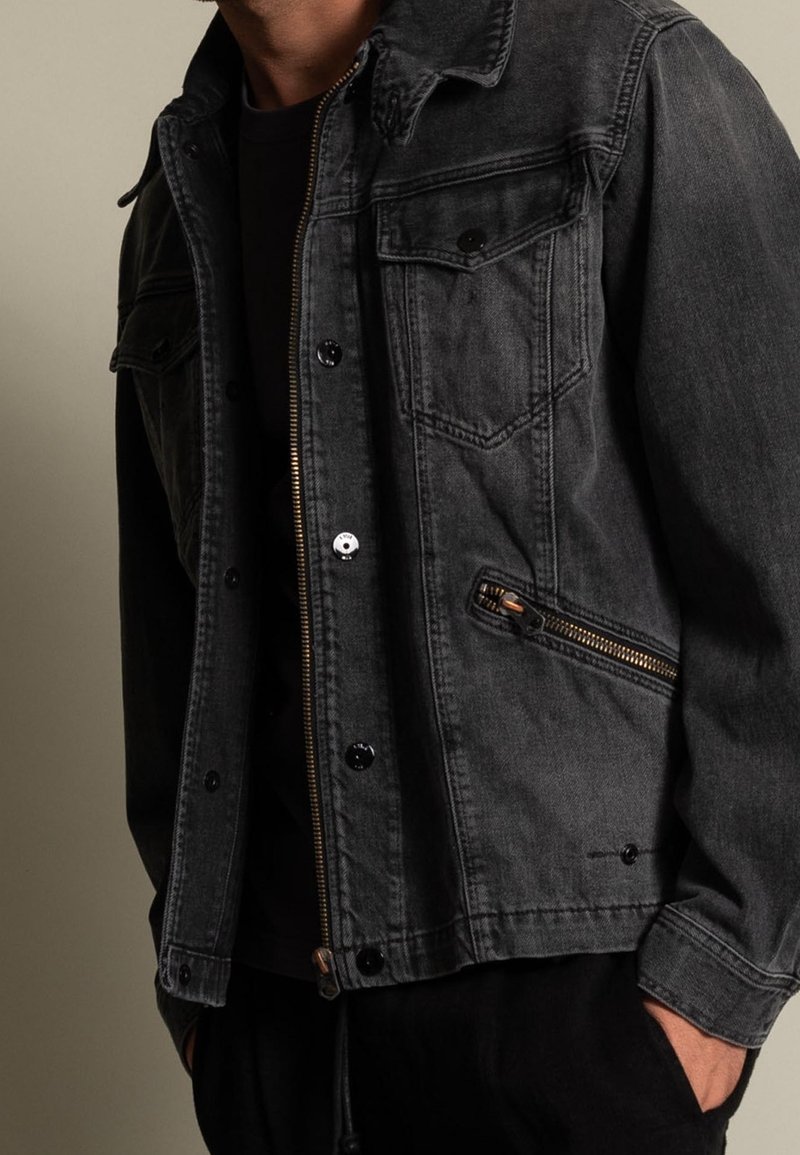 Man som bär en mörkgrå denimjacka med dragkedje- och knappfickor, svart skjorta under och händerna i fickorna på svarta byxor.