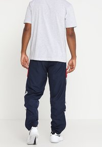 Námořnicky modré teplákové kalhoty s červenými akcenty a bílými pruhy, vyrobené z lehkého materiálu, s elastickými manžetami a pohodlným střihem.