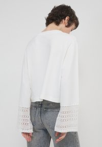 Top bianco a maniche lunghe cropped con dettagli in pizzo sulle maniche a campana, abbinato a jeans grigi a vita alta, visto da dietro.