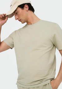 Lys beige kortærmet t-shirt med et struktureret, vaffel-lignende mønster. Modellen bærer en beige kasket og justerer den med den ene hånd.
