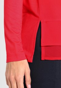 Red long-sleeve top with a slightly loose fit, smooth texture, side slit detail, and stitched seams, paired with black textured pants.