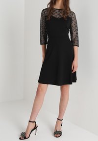 Svart klänning med trekvarts genomskinliga ärmar som har ett geometriskt mönster, utsvängd kjol och en figurnära midja. Bärs med svarta klacksandaler.