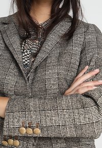 Vlněný blejzr v černo-bílém károvaném vzoru, s třemi kulatými hnědými knoflíky na každém rukávu; obsahuje klopu s notch a padnoucí střih.