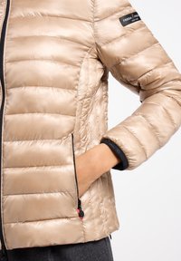Beżowa pikowana kurtka puchowa z zamkiem błyskawicznym z przodu, boczną kieszenią i czarnym mankietem. Posiada subtelną połyskliwość i znak firmowy na rękawie.