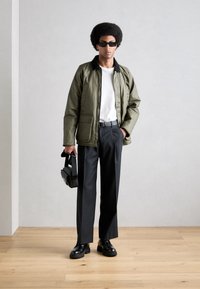 Chaqueta acolchada verde oliva con detalles negros, camiseta de cuello redondo blanca, pantalones negros y zapatos gruesos negros. Modelo sosteniendo un pequeño bolso negro.